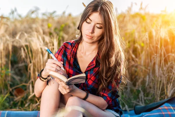 Chica sentada en una camisa de campo, trigo relajante en la naturaleza, hermoso cabello moreno. Escribe ideas en un cuaderno. Un estudiante después de la escuela. Notas conceptuales . —  Fotos de Stock