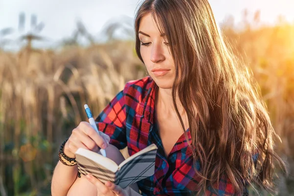 Chica sentada en una camisa de campo, trigo relajante en la naturaleza, hermoso cabello moreno. Escribe un bloc de notas de la idea. Un estudiante después de la escuela. Concepto de planes para el futuro . —  Fotos de Stock