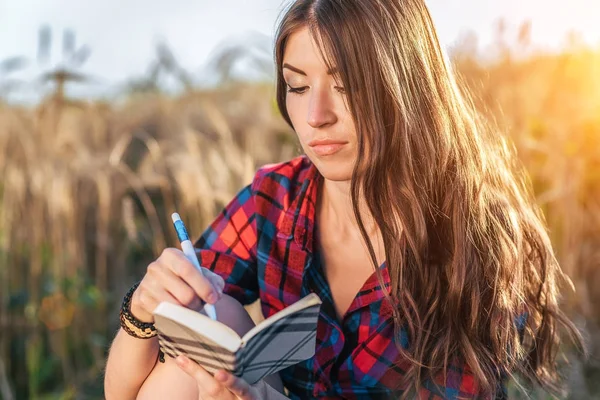 Meisje zit in een veld shirt, tarwe ontspannen in de natuur, mooie Donkerbruine haren. Schrijft een Kladblok voor het idee. Een student na school. Concept van de plannen voor de toekomst. — Stockfoto