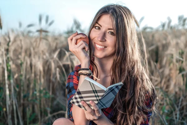 Ragazza seduta in una camicia da campo, frumento rilassante nella natura, bei capelli castani. Possiede una mela, un blocco note per il futuro. Felice sorridente guardando in cornice . — Foto Stock