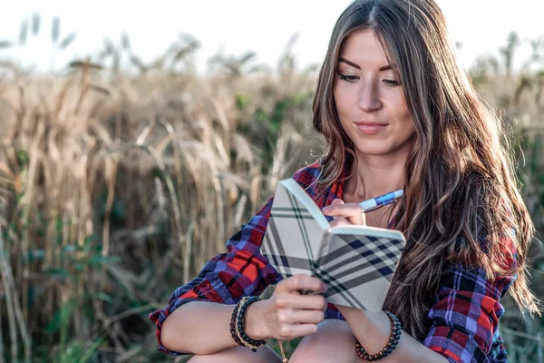 Девушка сидит в футболке, пшеница расслабляется на природе, красивые брюнетки. Хранит записную книжку на будущее. После уроков он пишет задание. Концептуальные записки являются уроками для — стоковое фото