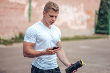 Bir adam bir shaker, bir kasa, bir protein bir şişe bir mesaj telefonda, sosyal ağlar, eldeki okur. Şehirde yaz aylarında. Sağlıklı bir yaşam tarzı kavramı.