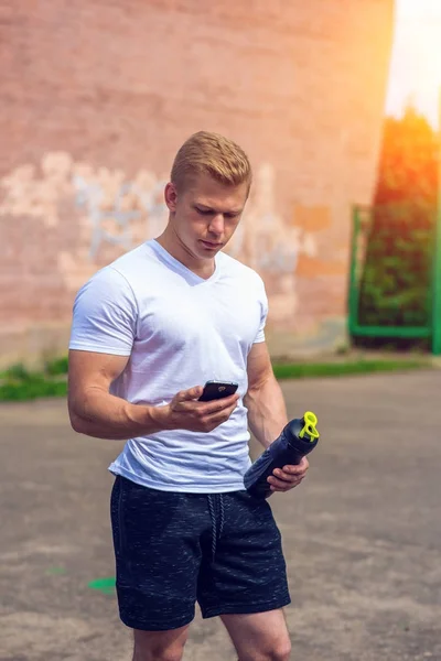Ein Mann liest eine Nachricht am Telefon, soziale Netzwerke, die Hand einen Shaker, eine Flasche Tresor, ein Protein. im Sommer die Stadt. das Konzept eines gesunden Lebensstils. im Smartphone Video ansehen. — Stockfoto