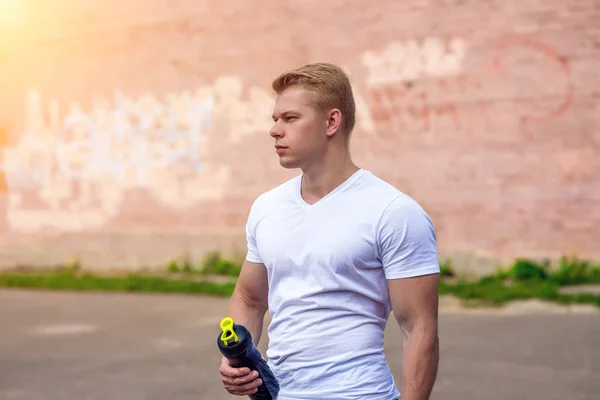 Um atleta na natureza, depois de treinar, em sua mão uma garrafa de água, uma coqueteleira com uma proteína. O ônibus controla o treinamento na cidade . — Fotografia de Stock