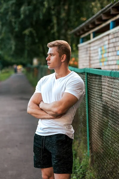 Der Mann steht im Sommer im Park, der Trainer schaut in die Ferne, in weißem T-Shirt und schwarzer Shorts. Lifestyle, ein gesunder, aufgepumpter Bodybuilder. — Stockfoto