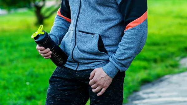 Close-up of a man holding a water bottle of protein in a sports suit in a park in the open air in the autumn of spring. — Stock Photo, Image
