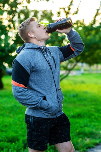 Athlete man listening to music on headphones. Drinks a bottle of water protein. Lifestyle athlete. Healthy eating. In the summer in the park.