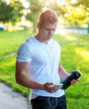 Vücut geliştirmeci Koç su elinde bir şişe ile kulaklık smartphone video izliyor. Smartphone müzik dinler. Kaslı bir atlet, doğada bir yaşam tarzı.