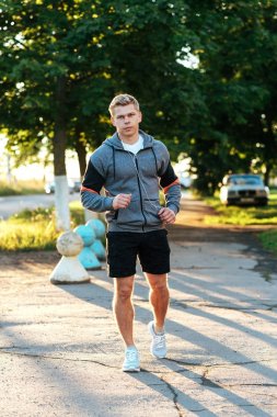 Bir erkek eğitmen kulaklıklar giyen yolun aşağısında çalışıyor. Parkta koşucu sabah. Müzik, kendinden emin bir görünüm dinliyor. Yaz yaşam tarzı, motivasyon güçlüdür. Açık havada şehirde.