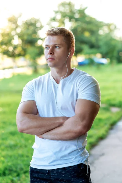 The man atlen stands in the park with headphones, a white T-shirt. Summer lifestyle, motivation is strong. Muscular arms. — Stock Photo, Image