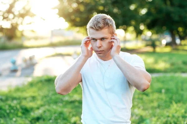 Athletiktrainer mit Kopfhörern. in weißem T-Shirt. Hörbuch, selbstbewusster Blick. Sommerlicher Lebensstil, Motivation ist stark. Draußen im Park. — Stockfoto