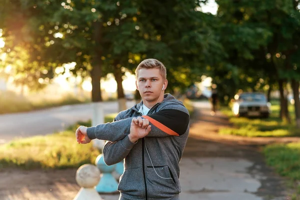 Ein männlicher Trainer knetet seine Hände vor dem Training mit Kopfhörern. Läufer am Morgen im Park. hört Musik, sieht selbstbewusst aus. Sommerlicher Lebensstil, Motivation ist stark. Draußen in der Stadt. — Stockfoto