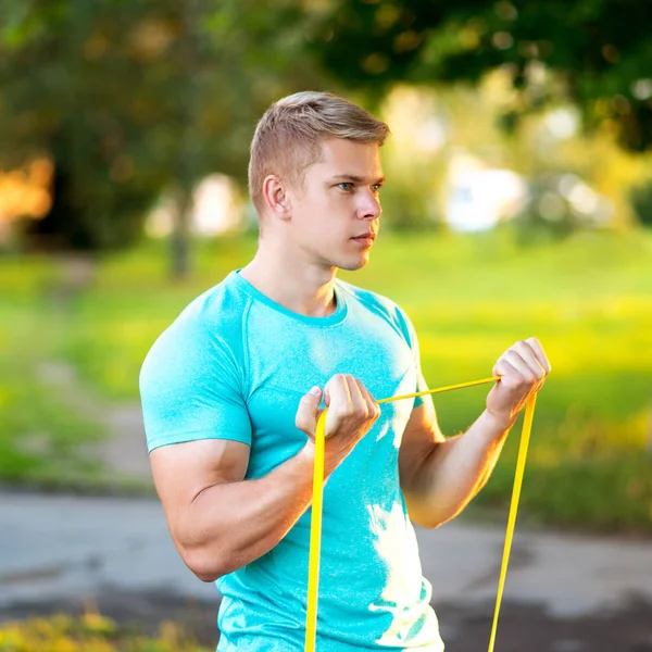 Sportovec v tričku třese svaly s školení smyčky s elastickým pásem. Letní životní styl, je silná motivace. Venku v parku. — Stock fotografie