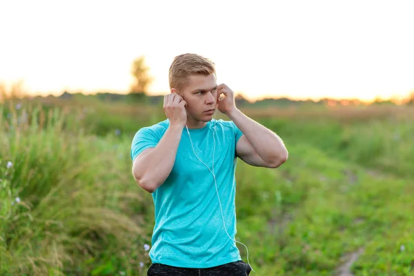 Ein mann atlen steht im park kleidet die kopfhörer, selbstbewussten blick. Sommerlicher Lebensstil, Motivation ist stark. Strickjacke mit T-Shirt. — Stockfoto