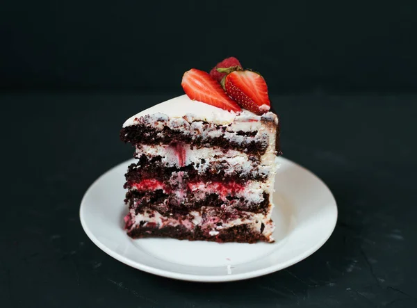 A slice of cream cake. On a black background and a white saucer, with strawberries. Dietary white from cream, with chocolate layers of berries. — Stock Photo, Image