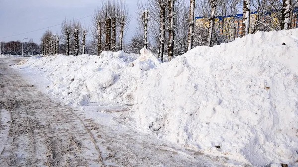 Уборка снега зимой в городе. Большие груды снега, собранные автомобилями на обочине дороги. Очищенный асфальт для автомобилей. На фоне высоких зданий и деревьев . — стоковое фото