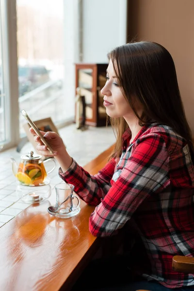 A female student in a shirt, in the afternoon at a cafe by the window, looks into the phone, looks at social networks. — Stock Photo, Image