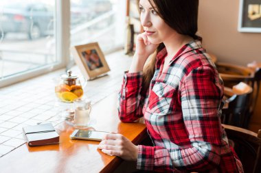 Bir kız öğrencinin gömlekli, gün boyu pencerenin, bir kafede pencereden görünüyor, yanındaki telefon mübarek çaydanlık ve bir kupa.