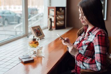 Bir kız öğrencinin gömlekli, öğleden sonra pencerenin görünüyor telefona, bir kafede bir ileti, bir demlik çay, ahşap bir masa üstünde bir defter bir kupa ile yazar.