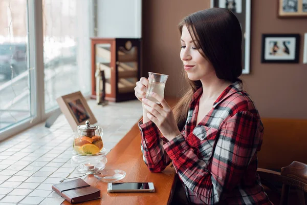 Una estudiante con una camisa sonríe felizmente, durante el día en un café junto a la ventana, sueña y sueña, sostiene una taza de té en sus manos, junto a un número de teléfono y una tetera . —  Fotos de Stock