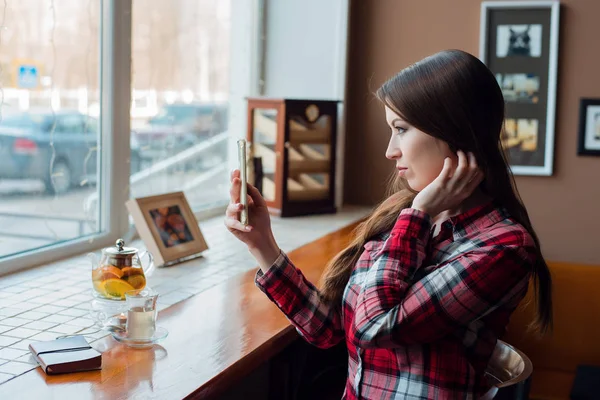 Студент дівчини в сорочці, вдень в кафе біля вікна, дивиться в телефон, робить селфі, чайник з чашкою чаю, блокнот на дерев'яному столі . — стокове фото