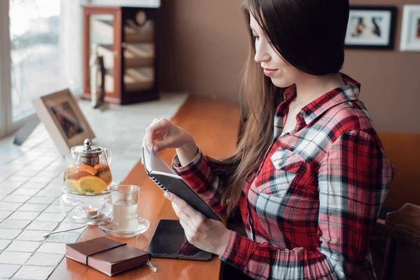 Студент дівчини в сорочці, вдень у кафе біля вікна, читає блок, чайник з чашкою чаю, сидить за дерев'яною стійкою. Планшет лежить . — стокове фото