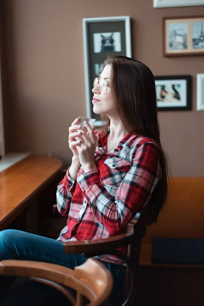 那个穿着眼镜和衬衫的黑发女孩, 下午在窗边的咖啡馆里, 早晨喝茶。拿着杯子喝一杯. — 图库照片