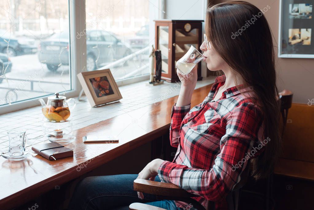 The brunette girl with glasses and a shirt, in the afternoon at a cafe by the window, drinks tea in the morning. Next to the table there is a kettle with a telephone and a paper pad.