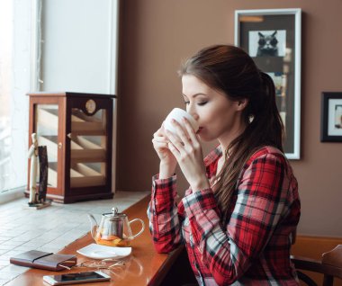 Kız öğrenci ekose gömlekli, öğleden sonra bir masada, bir bardak çay, bir kafede kahve içme. Bir smartphone gözlük ve kağıt defter ile geçerli. Esmer kadın.