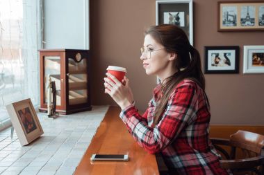 Kız öğrenci bir ekose gömlek ve gözlük, öğleden sonra kırmızı bir fincan çay ile pencerenin yanındaki bir masada bir kafede. Sıcak kahve elinde tutar. Esmer bir kadın pencereden görünüyor.