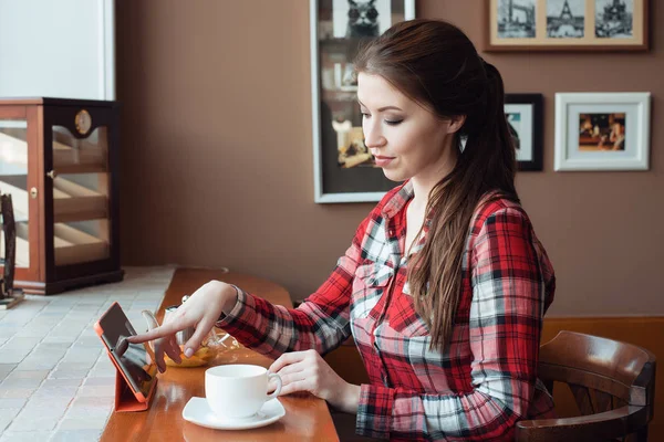 Студентка в клетчатой рубашке, днем в кафе за столом, с кружкой чая. На столе планшет, выбирает на сенсорном экране. Брюнетка делает заказ в Интернете . — стоковое фото