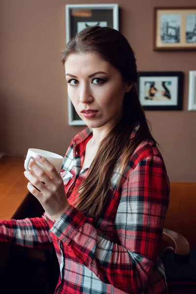 Žena v košili. Brunetka s dlouhými vlasy. V rukou šálek kávy nebo čaje. Pózování s překvapen emoce na tváři. Odpoledne v kavárně u okna. — Stock fotografie