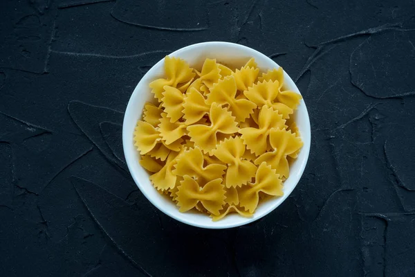Farfalle macaroni paste in a white cup on a black textured background, in the center close-up from the top. — Stock Photo, Image