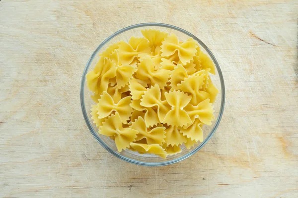 Farfalle macaroni pasta in a glass bowl on a wooden table texture background, in the center with the top. — Stock Photo, Image