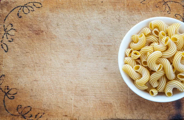 Macarrones de pasta rigati en un tazón blanco sobre una placa de madera de corte, fondo de textura con un lado. Primer plano con la parte superior. Espacio libre para texto . — Foto de Stock