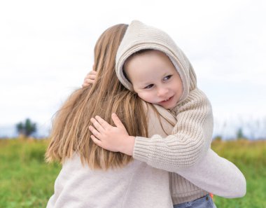 4-5 yaşlarında küçük bir çocuk, kapüşonlu sıcak süveter giymiş annesine sarılıyor, mutlu bir gülümsemeyle. Sonbahar parkında. Aşk duyguları, bakım anlayış, güven ve destek..