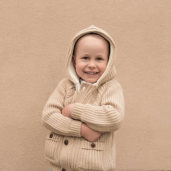 4-5 살의 남자 아이가 후드를 쓰고 행복하게 웃는 따뜻 한 스웨터를 입고 벽에 붙어 있습니다. 긍정적 인 기분 과 새해 기분의 기쁨. — 스톡 사진