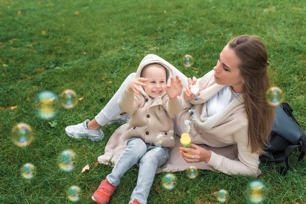 Matka žena s malým chlapcem, syn 4-5 let, ležet na trávě. Mýdlové bubliny jsou vypuštěny. Šťastní lidé hrají smích a baví pozitivní emoce, v teplém oblečení na podzim, ve svetru s kapucí. — Stock fotografie