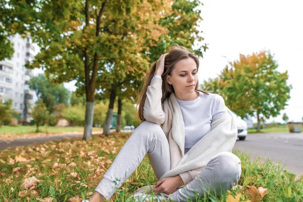 Красивая и молодая женщина сидит в осенний день в парке на дороге, фоновые деревья и листья деревьев. Отдыхает на природе. Эмоции комфорта и заботы . — стоковое фото