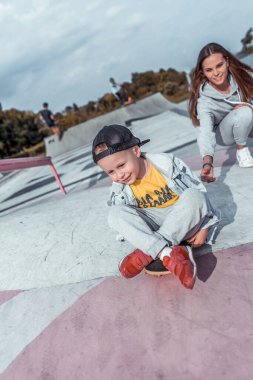 4-5 yaşlarında küçük bir oğlan çocuğu, genç bir anne kadın, kaykay sürmeyi öğreniyor, yazın şehir parkında oyun parkında. Kaykay, gündelik kıyafet. Denge eğitimine destek olun. Neşeli duygular eğlence.