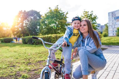 Mutlu bir gülüş, anne kadın ve 4-5 yaşlarında küçük bir oğlan çocuğu, bisiklet sürmeyi öğrenin, eğlenin ve şehir parkında yaz tatilinin tadını çıkarın. Günlük kot şapkası, spor ayakkabı pantolonu..