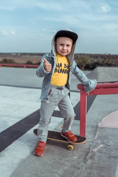 Anak laki-laki berusia 4-5 tahun, di musim gugur di kota di lapangan olahraga, belajar untuk naik skateboard, tersenyum bahagia, menunjukkan jari ke atas. Casual memakai topi denim, celana olahraga . — Stok Foto