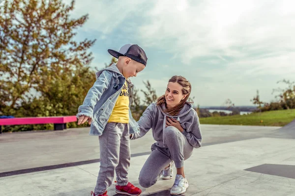 Happy mom woman with little boy son 4-5 years old, learns to ride skateboard, summer autumn in city park playground. Skateboard, casual wear. Support help and balance training. Emotions of joy fun. — Stockfoto
