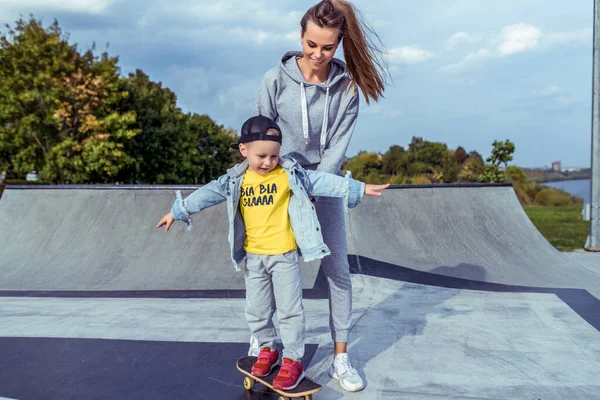 Ibu wanita anak laki-laki yang bahagia 4-5 tahun, belajar untuk naik skateboard, musim panas musim gugur di taman bermain kota. Skateboard, pakaian kasual. Dukungan untuk keseimbangan latihan. Emosi sukacita menyenangkan . — Stok Foto