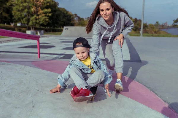 Ibu muda wanita dengan anak laki-laki berusia 4-5 tahun, belajar naik skateboard, musim panas musim gugur di taman bermain kota. Skateboard, pakaian kasual. Dukungan untuk keseimbangan latihan. Emosi sukacita menyenangkan . — Stok Foto