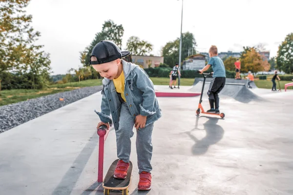 Little boy 4-5 years old, autumn summer in city on sports field, learning to ride skateboard, happy smiling, free space text. Casual wear denim cap, sneaker pants. Background kids teens. — Stockfoto