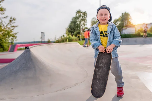 Happy little boy 4-5 years old, in autumn summer city on sports field, learning to ride skateboard, smiling, free space for text. Casual wear denim cap, sneaker pants. Background kids teens. — Stockfoto