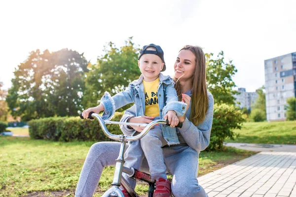 Mamá mujer con niño pequeño hijo de 4-5 años de edad, aprender a montar en bicicleta, divertirse riendo y disfrutar de sus vacaciones, otoño de verano en el sitio del parque de la ciudad. Casual use gorra de mezclilla, pantalones deportivos . — Foto de Stock