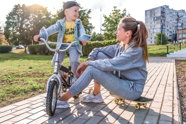 Selamat tertawa, Ibu wanita dengan anak laki-laki berusia 4-5 tahun, belajar mengendarai sepeda, bersenang-senang bersantai, musim panas jatuh di daerah taman kota. Pakai topi denim biasa, celana olahraga. Skateboard sedang duduk . — Stok Foto