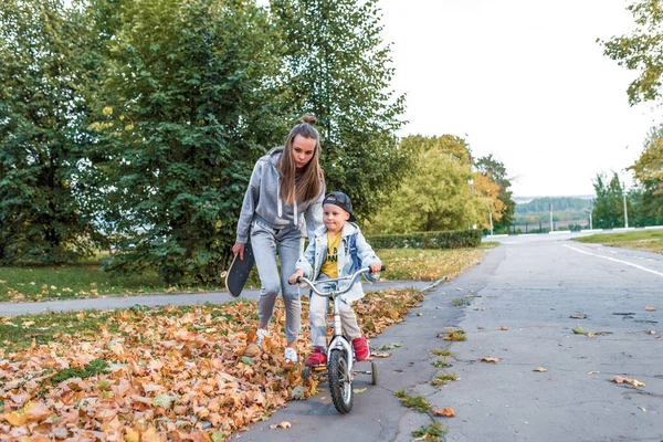 Mama femeie cu fiul băiețelului de 4-5 ani, învață să meargă cu bicicleta, să sprijine îngrijirea și educația, să echilibreze formarea, să se relaxeze în parcul de vară al orașului. Capac din denim, pantaloni de adidaşi. Ziua Mamei . — Fotografie, imagine de stoc